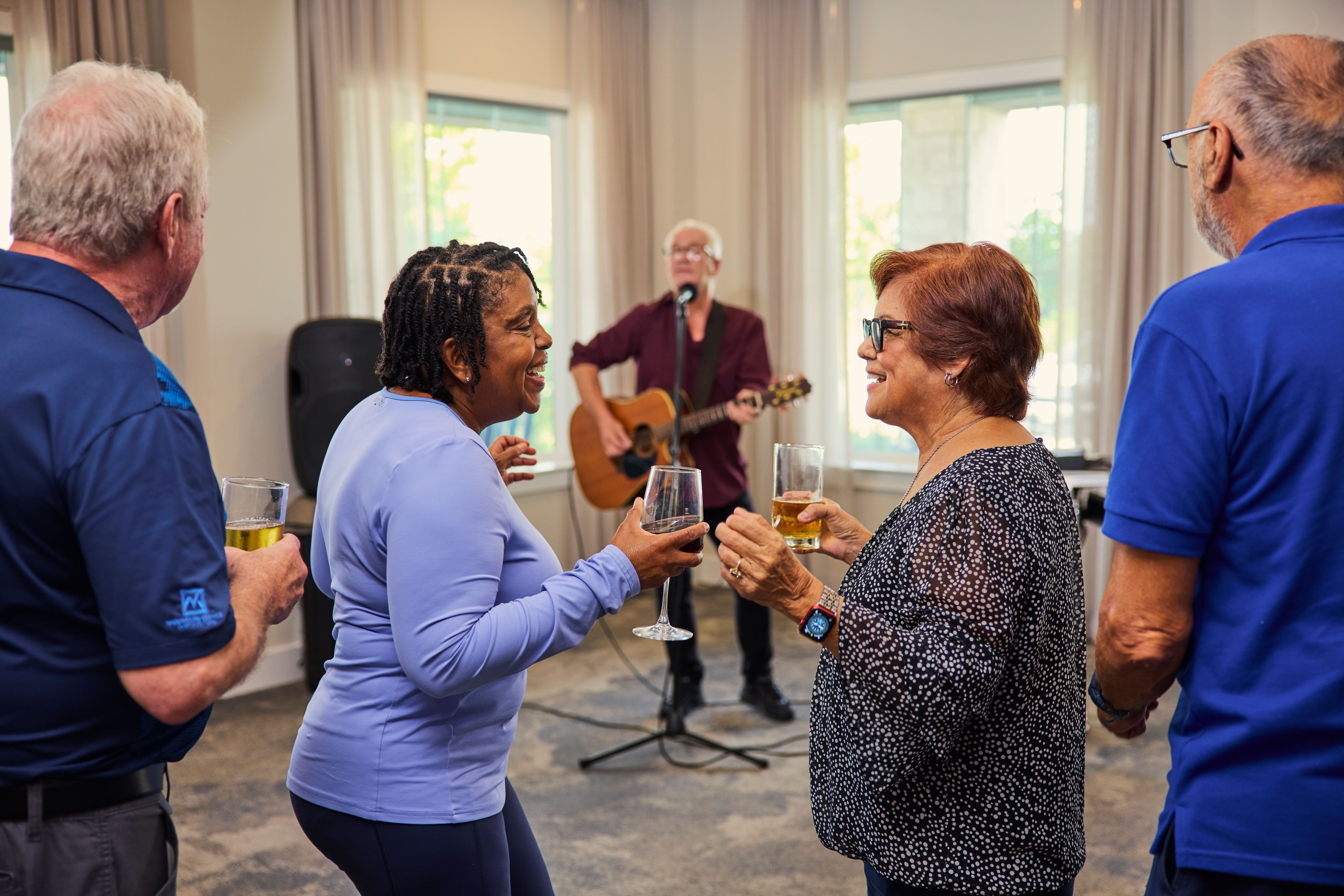 A group of people are gathered in a room, with one person playing a guitar and others holding glasses of wine.
